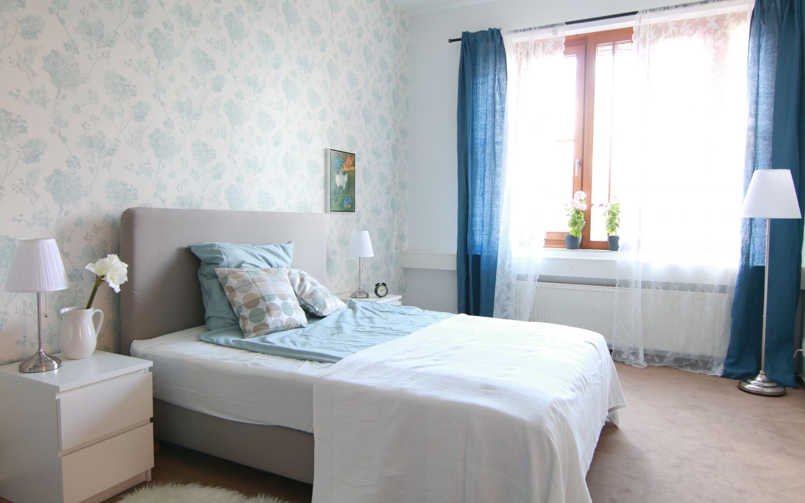 Graues Bett vor weiß-blauer Wand und hellen Möbeln. Dunkelblaue Vorhänge vor dem Fenster.